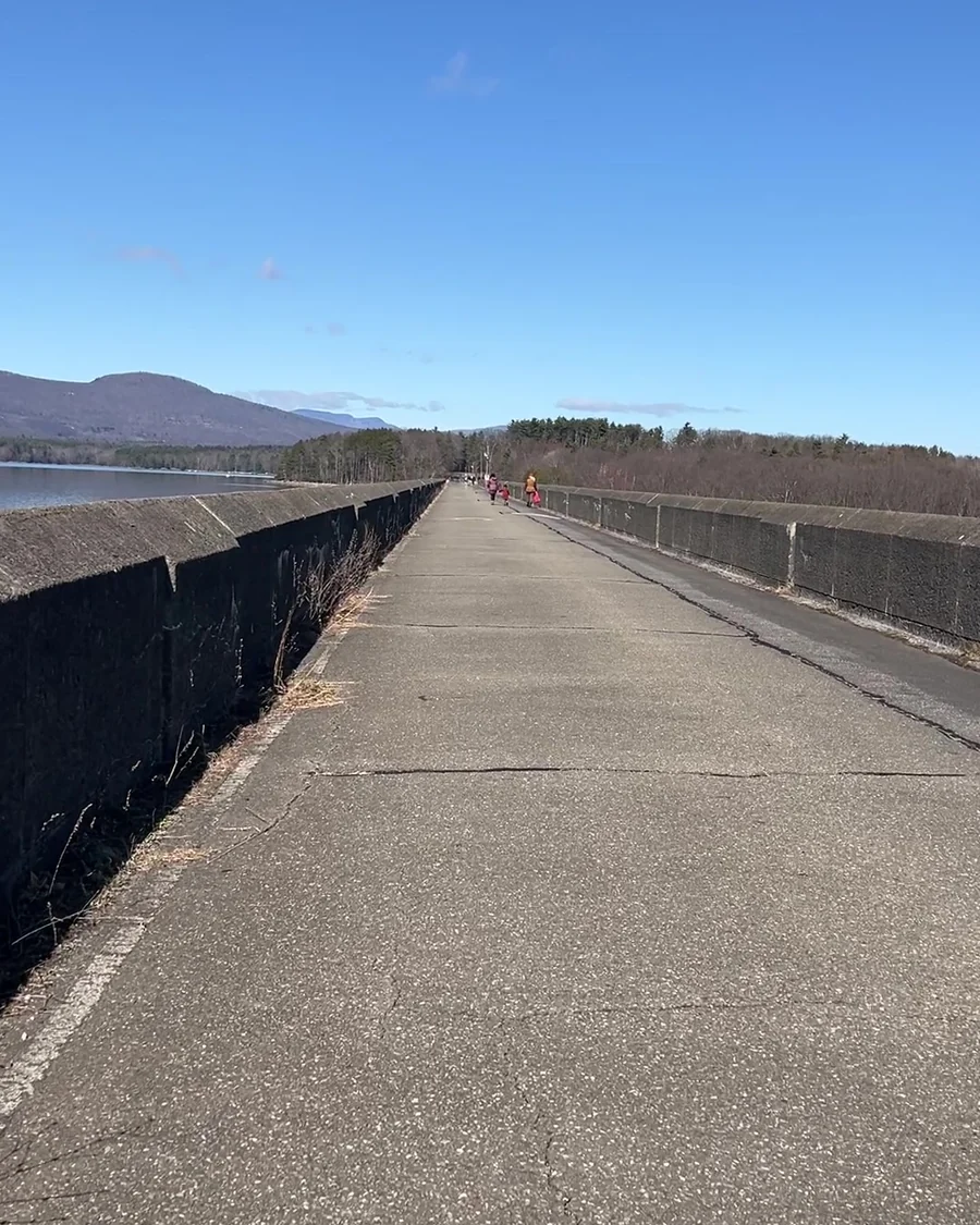 A paved walkway running straight out between low concrete walls toward the Ashokan Reservoir, with Catskill peaks across the water and a few distant walkers ahead