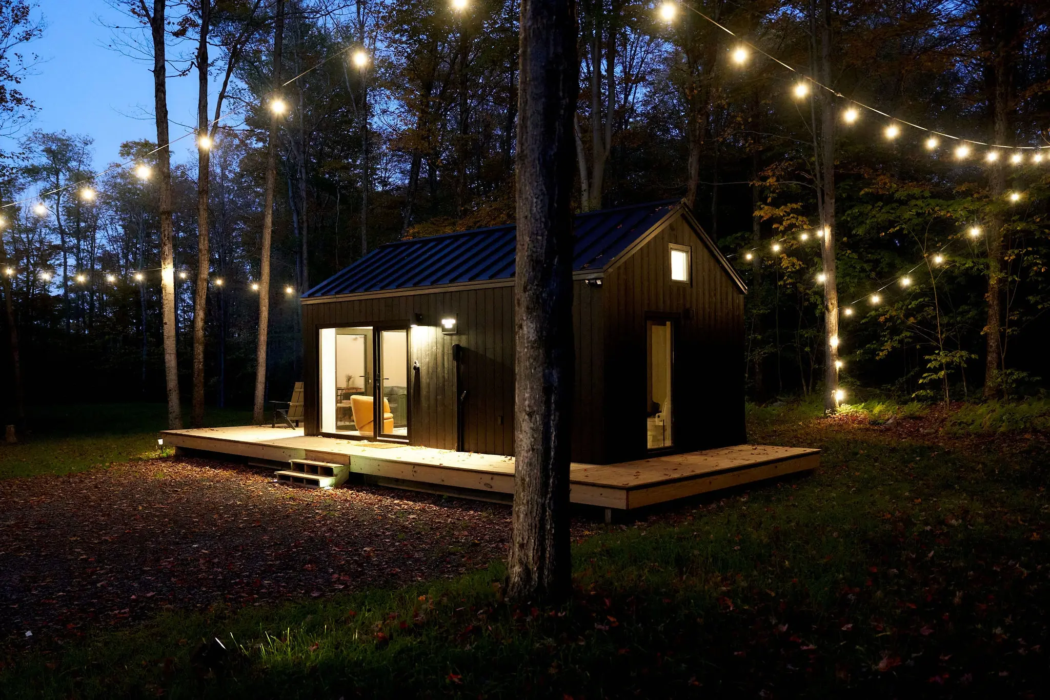 Small black cabin with glowing windows under string lights in the woods on a fall evening