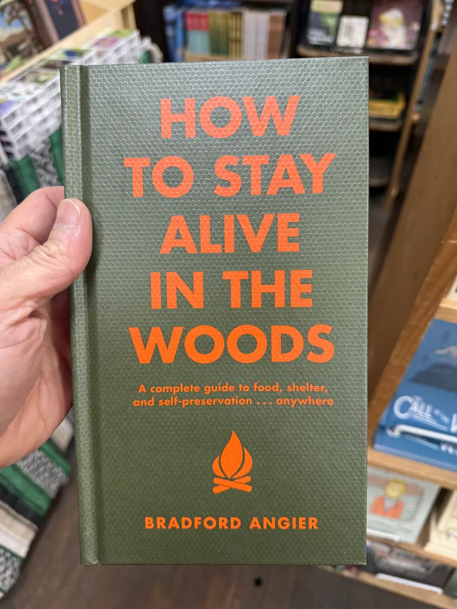 A hand holding a green hardcover copy of How to Stay Alive in the Woods by Bradford Angier in a Phoenicia bookshop