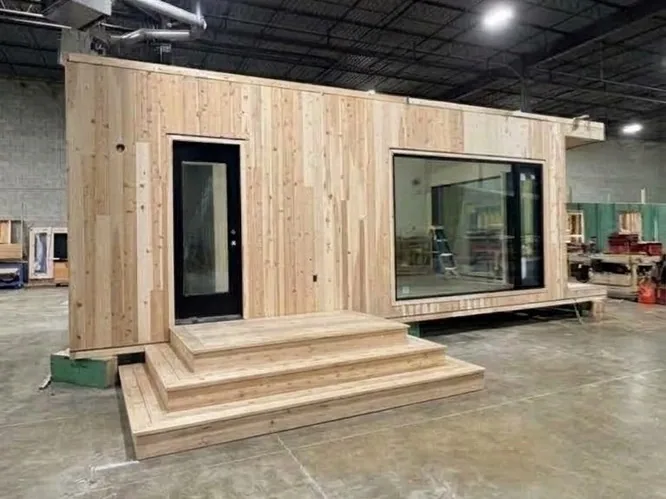 Cedar-clad tiny cabin studio with a black glass door, a large picture window and wide wooden steps, under construction inside a workshop