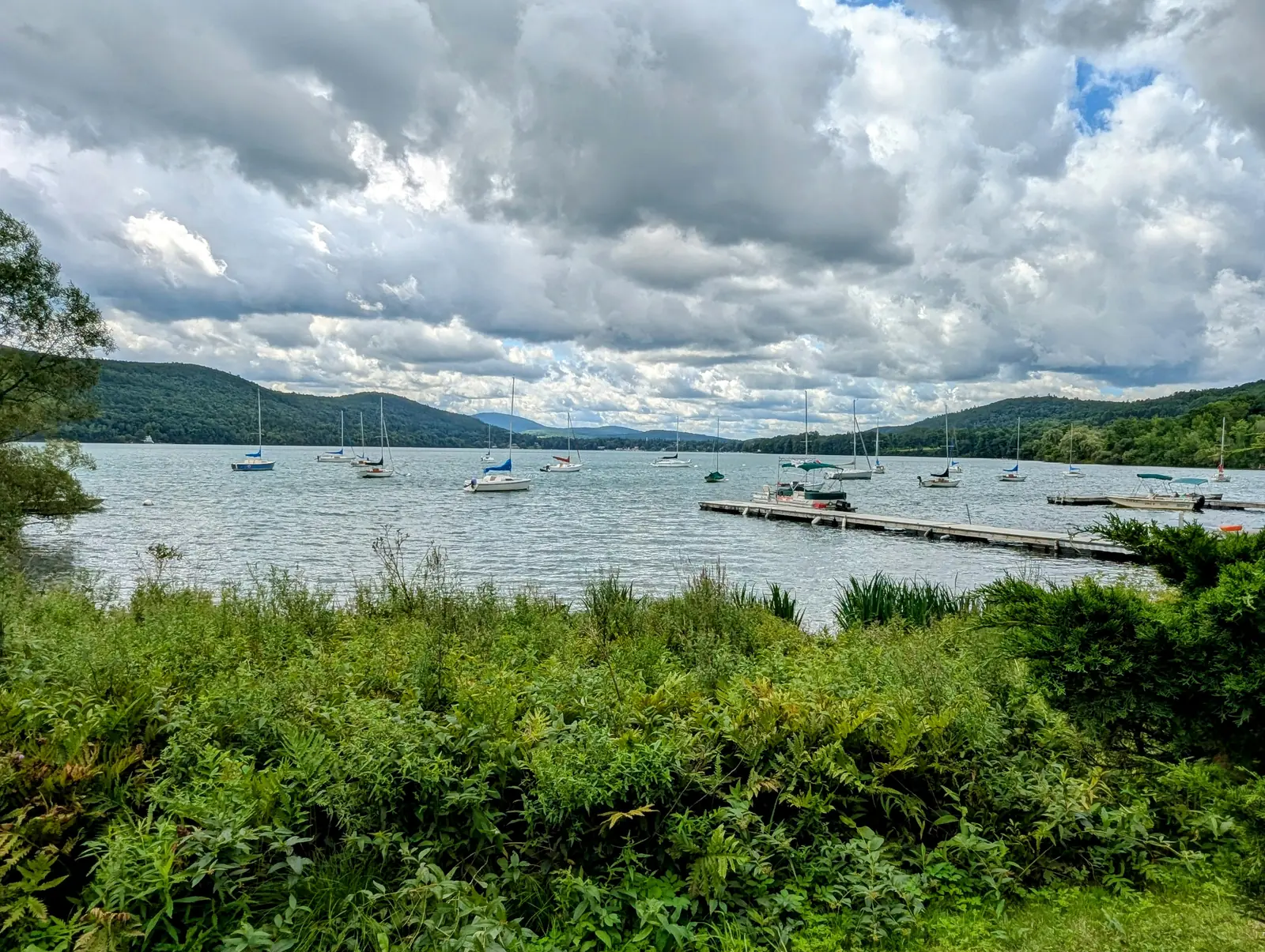 Sailboats moored on Otsego Lake below green hills, seen from the shore at Brookwood Point in Cooperstown, NY